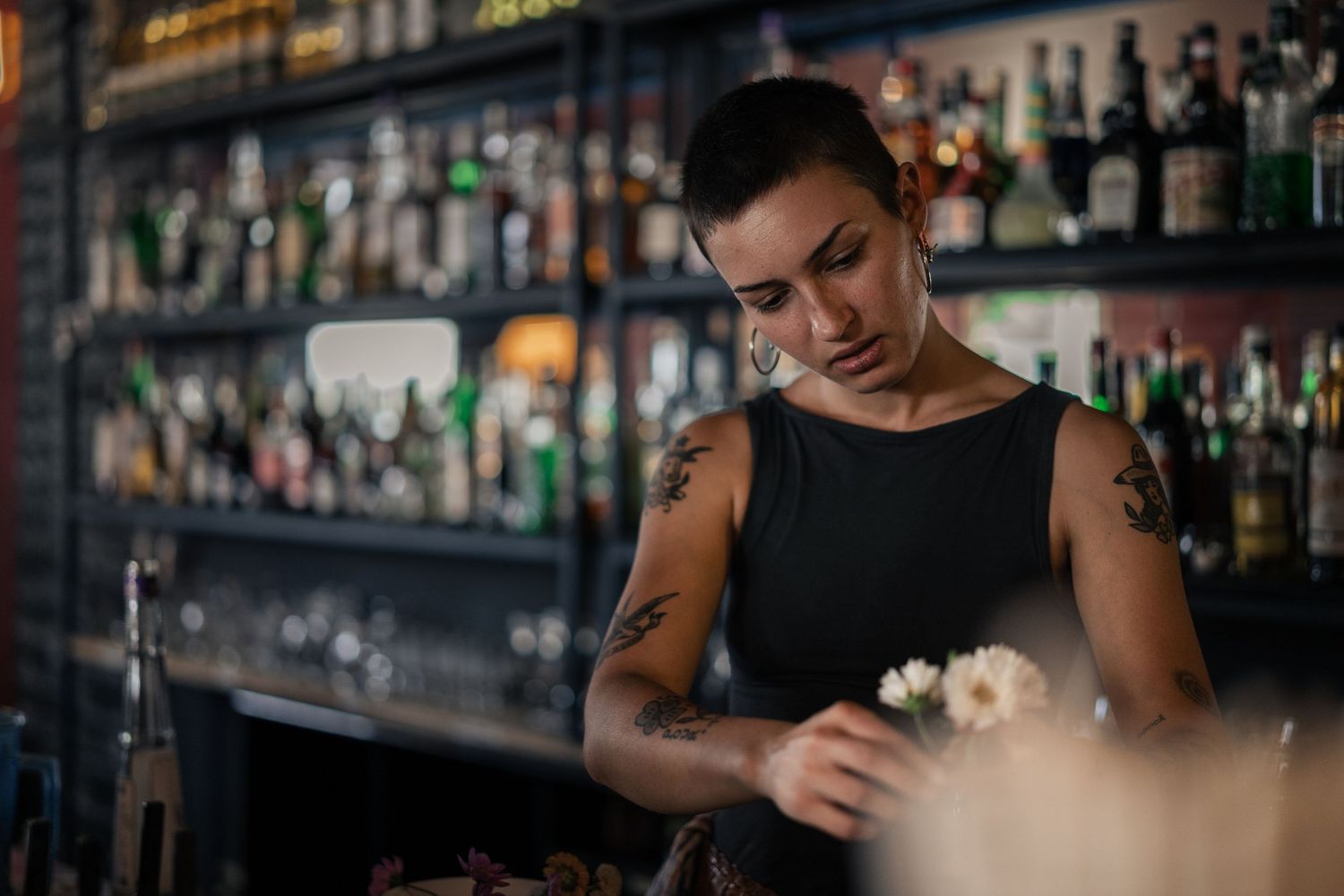 A tattooed female bartender carefully arranges flowers at a bar with bottles in the background.

Back Bar