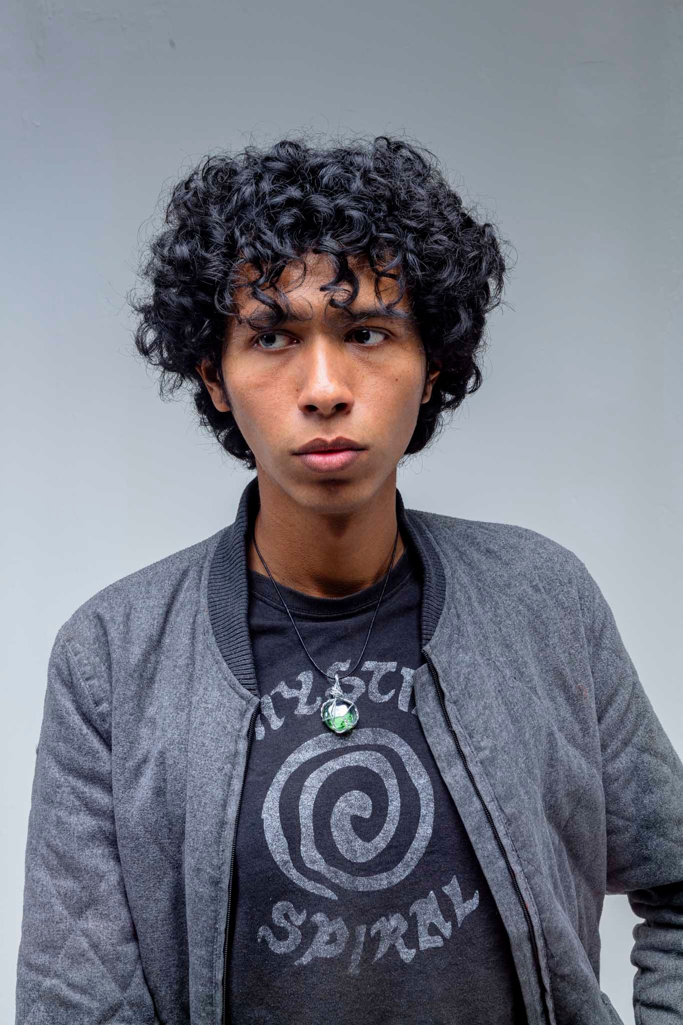 Young man with curly hair sitting on a stool wearing a gray jacket, jeans, and boots against a white background.