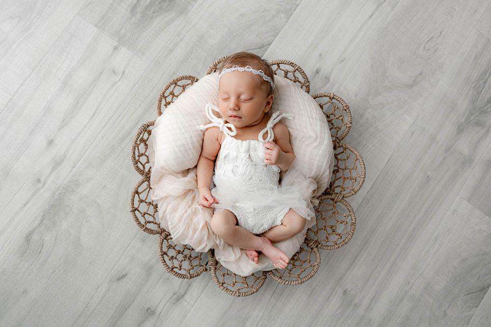 Sleeping newborn baby girl in white romper nestled in decorative floral basket with soft pink tulle.