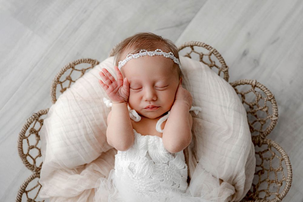 Sleeping newborn baby girl in white lace outfit and floral headband nestled in a wicker basket with cream blanket.