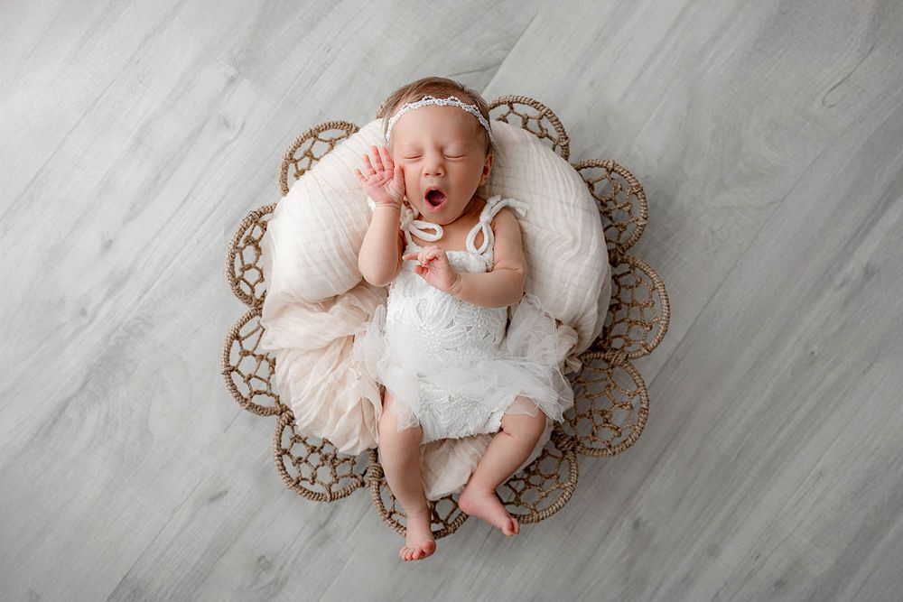 Yawning newborn baby girl in white tutu dress lying in wicker basket on soft cream pillow on gray floor.