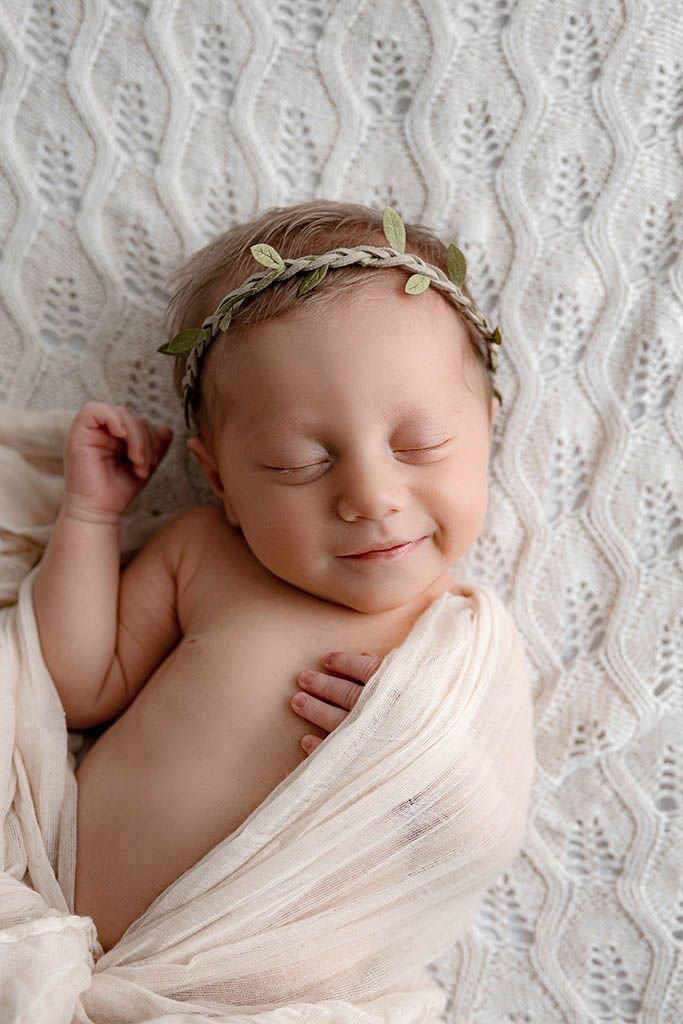 Smiling newborn baby wrapped in soft cream fabric, wearing a delicate green leaf crown, sleeping peacefully on a white knit blanket.