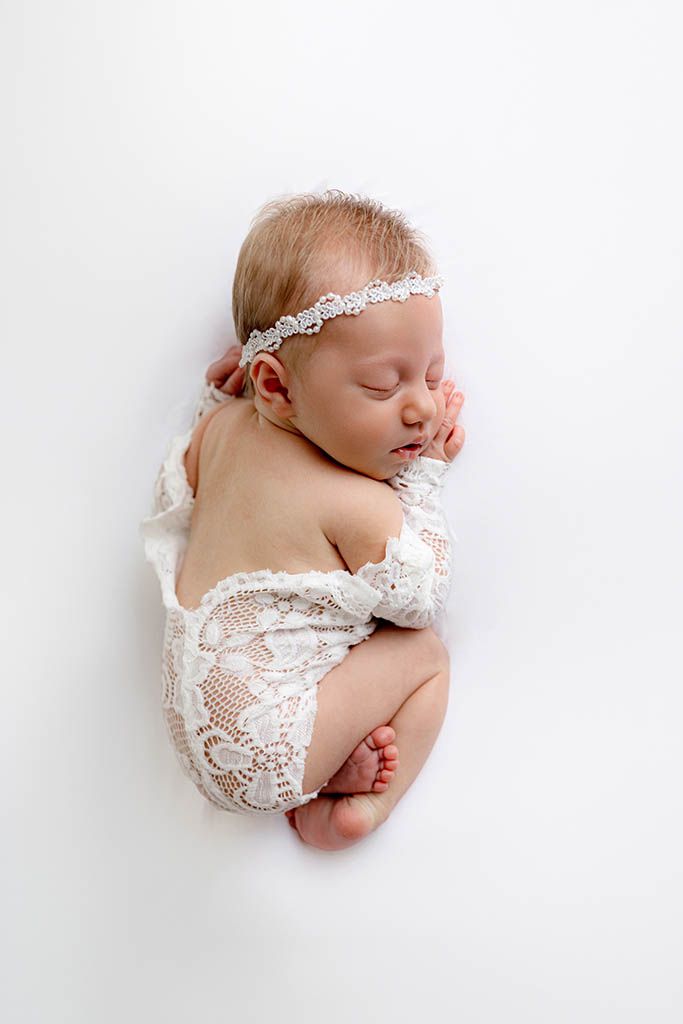 Sleeping newborn baby girl in white lace romper and floral headband, curled up on white background.
