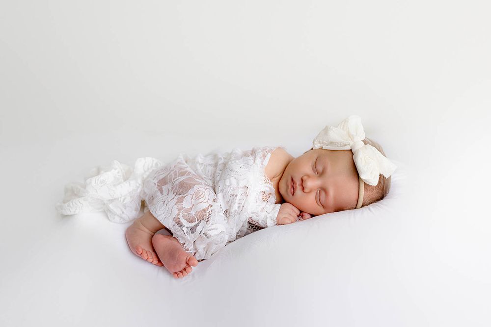 Sleeping newborn baby girl in white lace outfit and bow headband on white background.