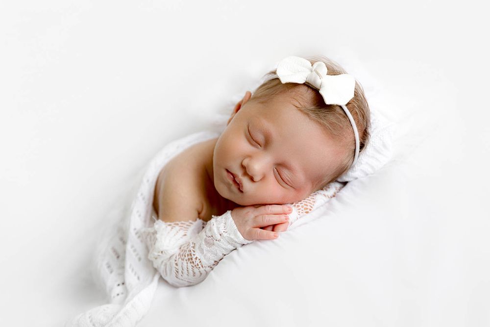 Peaceful newborn baby girl sleeping wrapped in white lace swaddle with white bow headband.