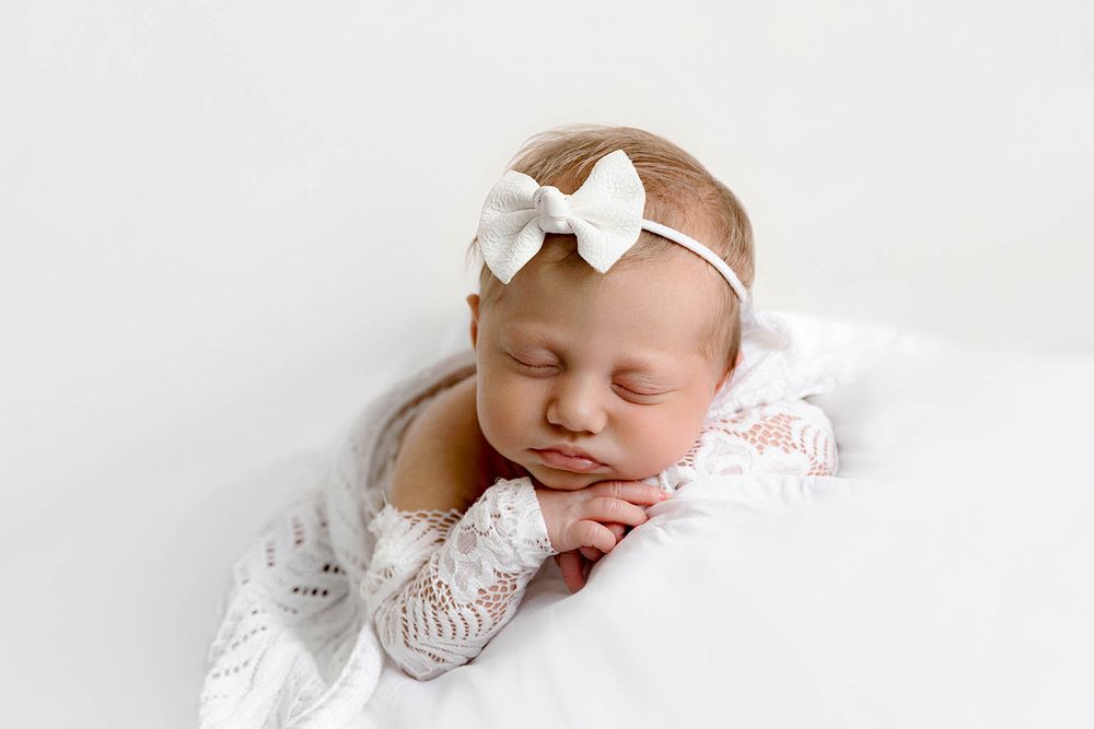 Sleeping newborn baby girl in white lace outfit and bow headband posed with hands under chin.
