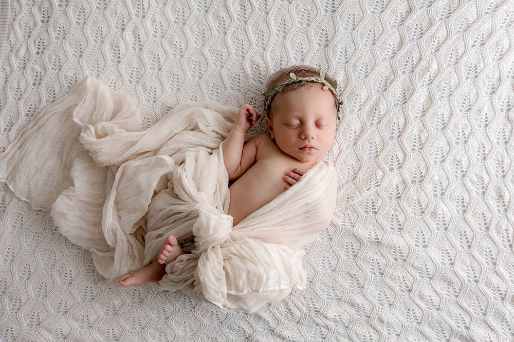 Sleeping newborn baby wrapped in cream gauze swaddle with floral headband on white knit blanket.