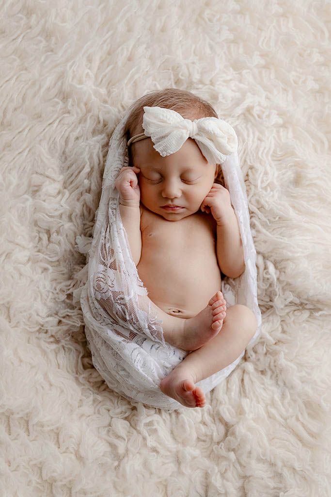 Sleeping newborn baby girl wrapped in white lace with a bow headband on a fluffy cream background.