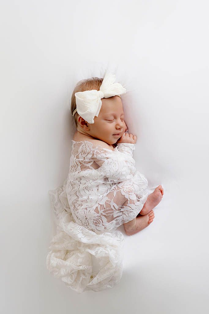 Newborn baby girl sleeping peacefully in white lace romper with large bow headband on white background.