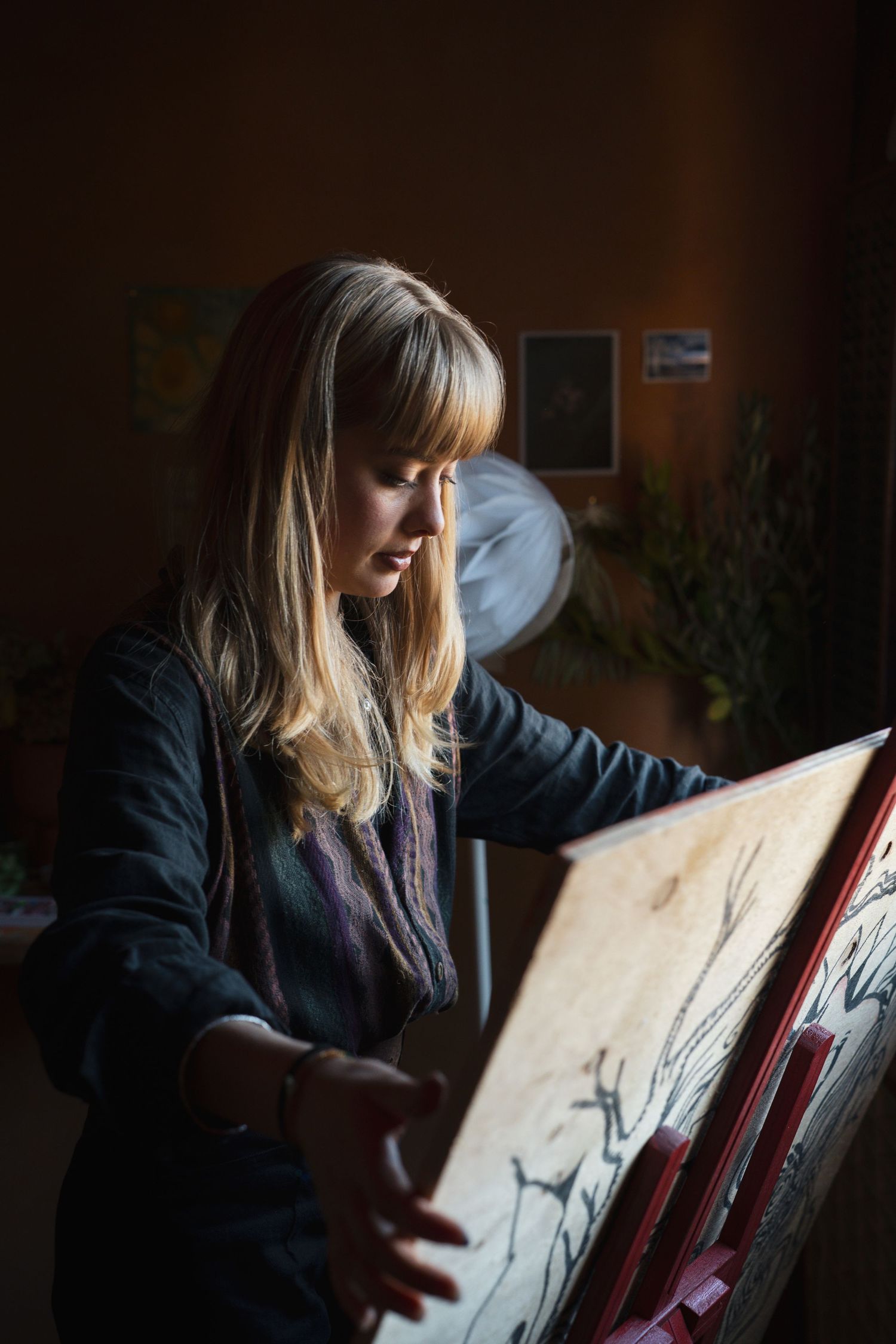 Female artist with blonde hair working on drawing at easel in dimly lit art studio with plants.

Maya Caroll-Tischler