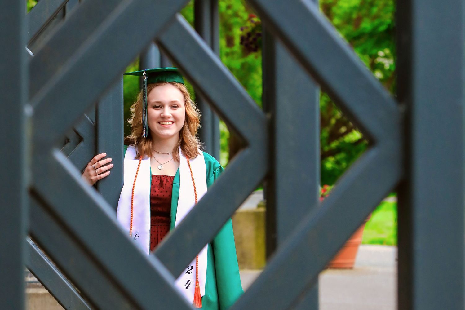 Graduate in green dress and white graduation stole poses behind decorative geometric architectural frame outdoors.