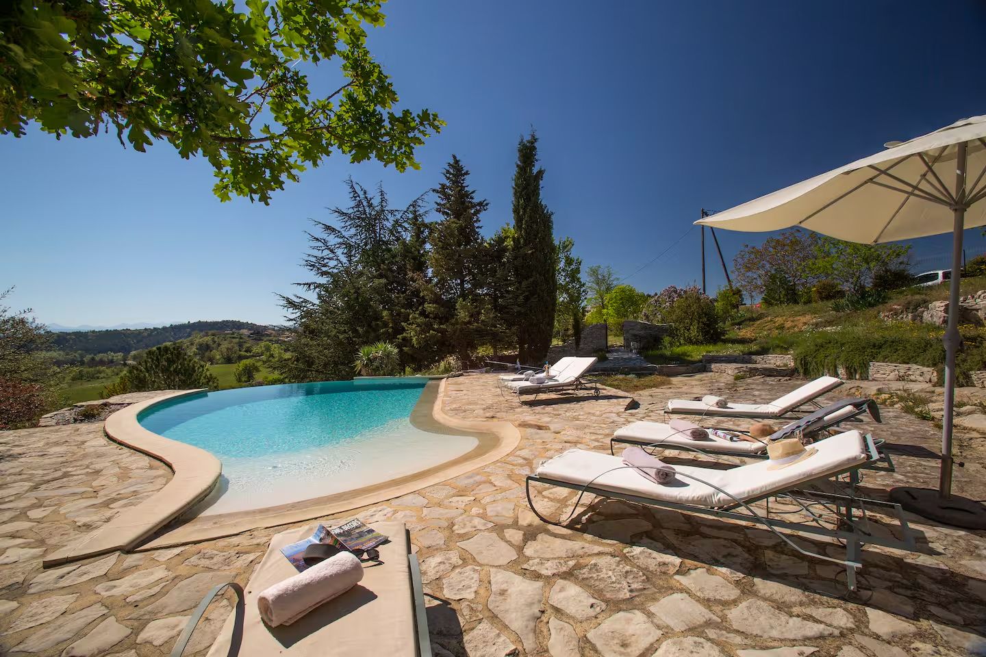 Curved swimming pool with lounge chairs on stone patio overlooking Tuscan countryside hills and cypress trees.