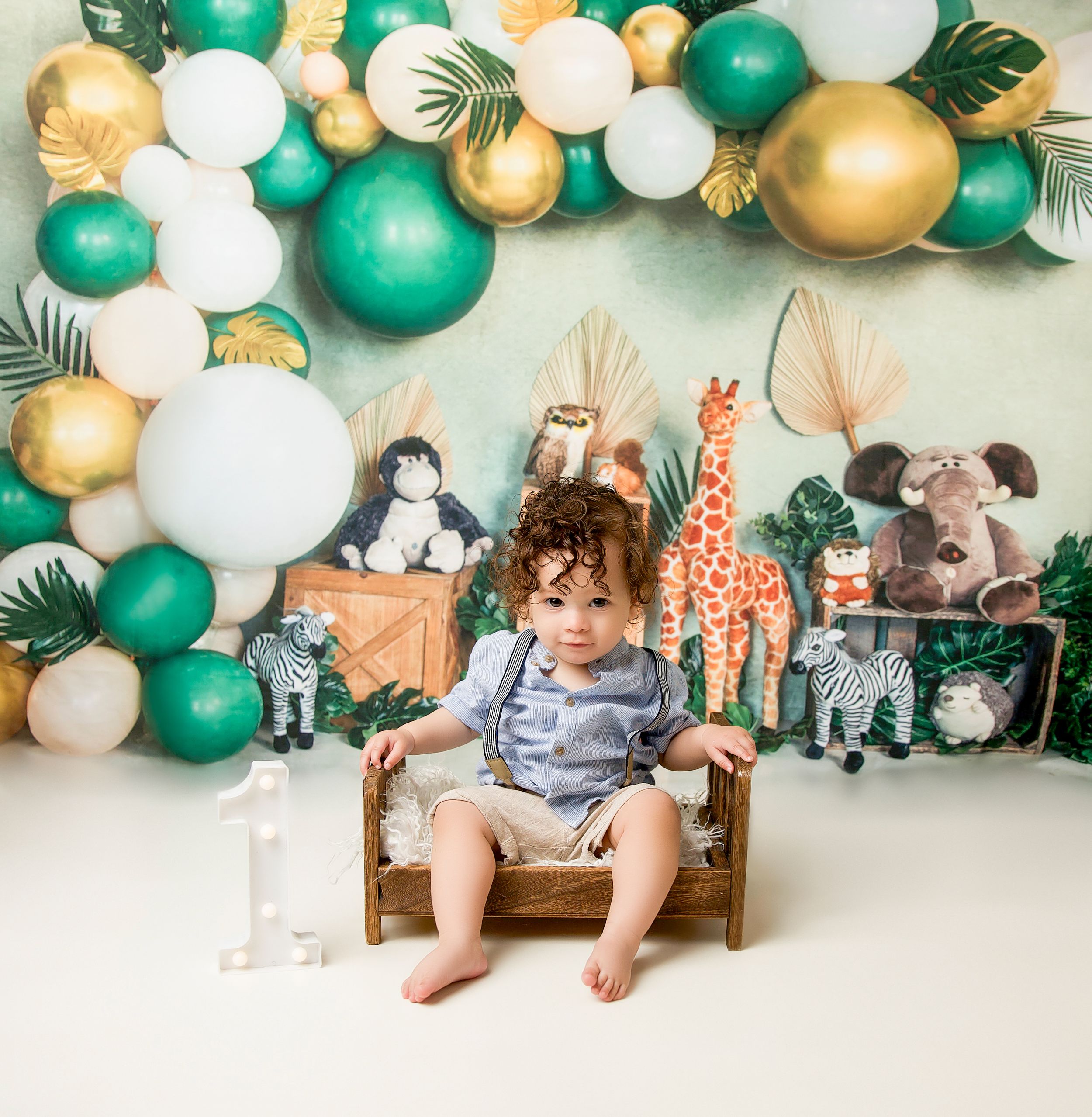 Safari themed party setup with green and gold balloon arch, decorative toy animals, and wooden chair against a white wall.