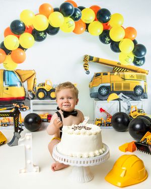 Construction themed first birthday party setup with yellow and black balloon arch, toy trucks, cake, and hard hat decor.