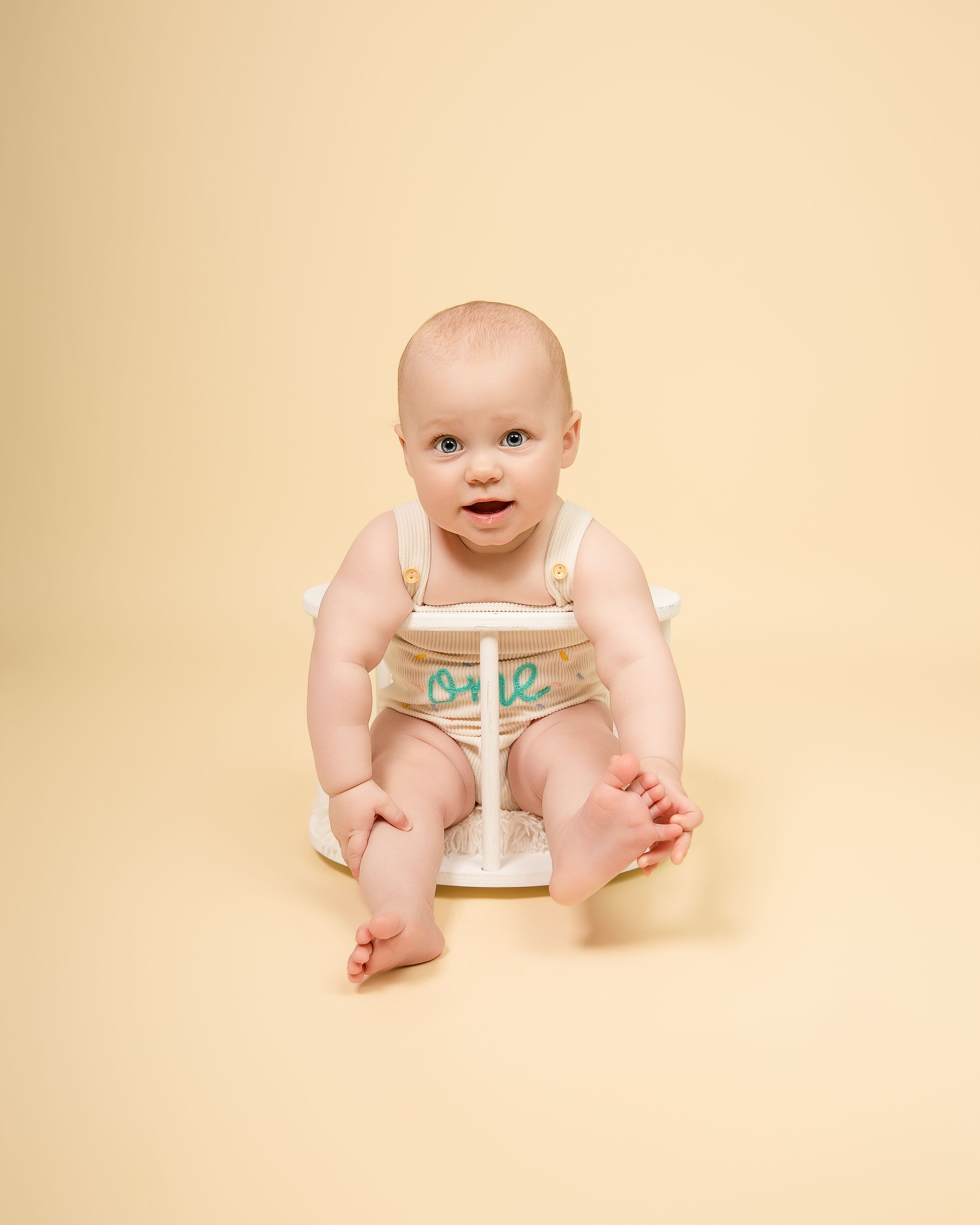 Adorable baby in a cream-colored diaper sits on a plain beige background looking up with wide eyes.