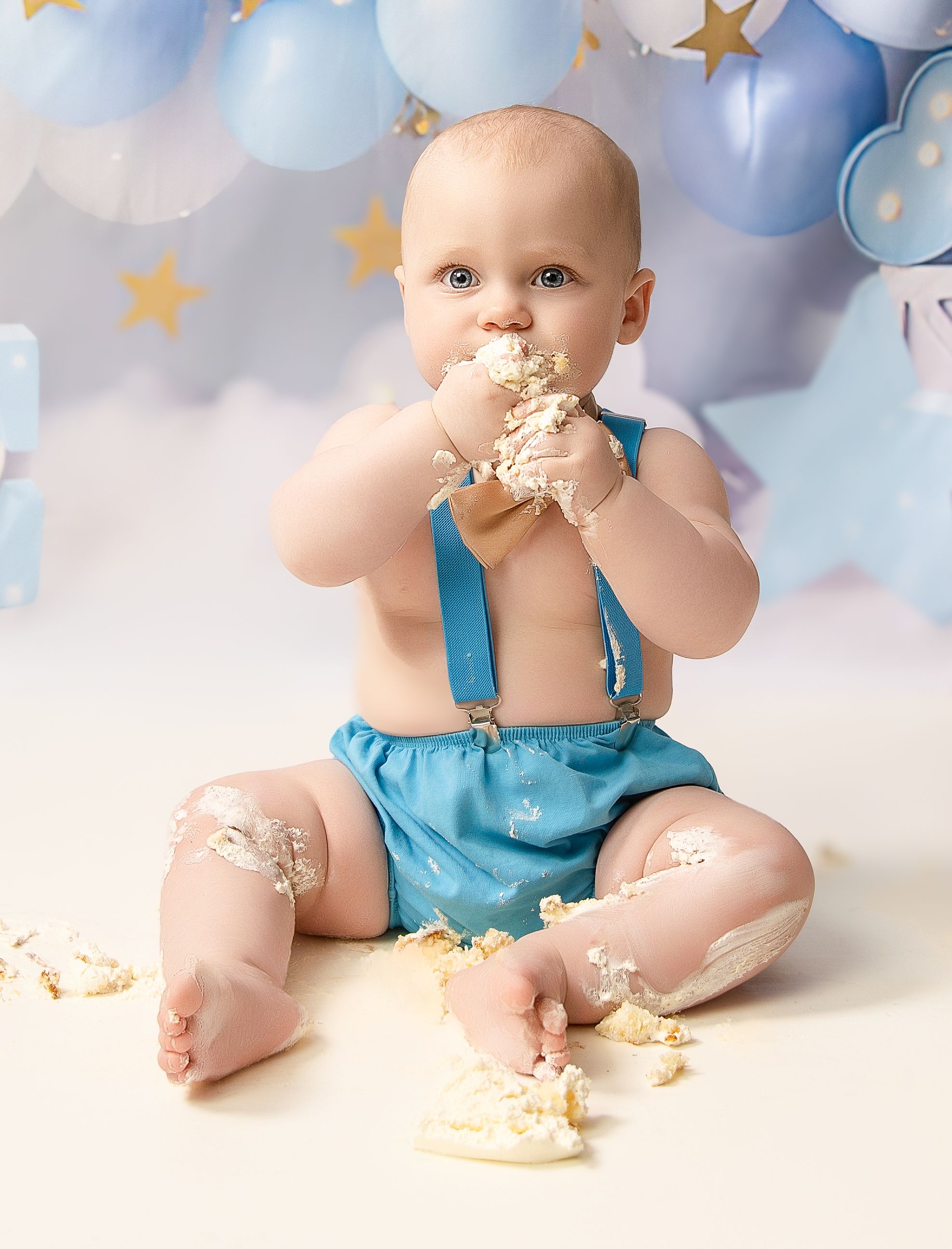 Baby in turquoise suspender shorts enjoying cake smash photo session with blue balloons and gold star backdrop.