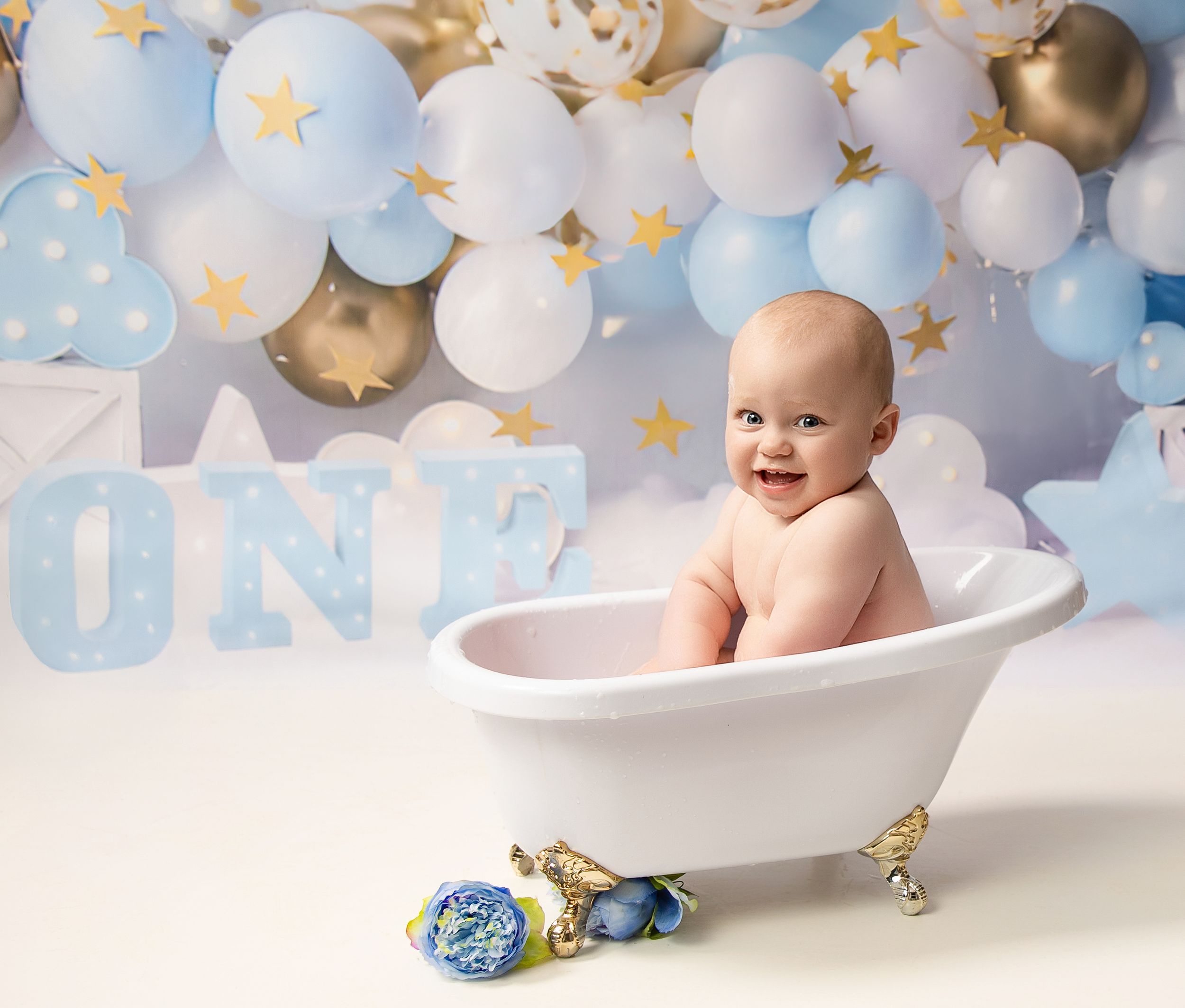 Baby sitting in miniature white bathtub with blue and white balloon backdrop and gold star decorations.