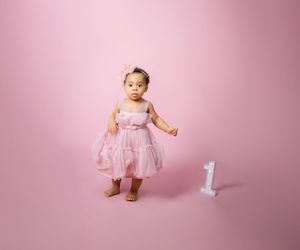 A baby in a pink tulle dress stands next to a number one candle against a pink studio backdrop.