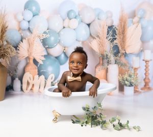 Baby wearing brown sweater smiling by blue and peach balloon arch with pampas grass and festive decorations for one year milestone.