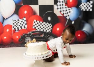 Racing-themed first birthday party setup with red and blue balloons, checkered flags, and white cake next to toy car on floor.