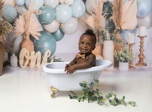 Festive cake smash setup with white bathtub, pink and blue balloon arch, pampas grass, and rustic decor.
