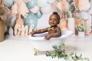 A small child sits in a decorated bathtub with teal and white balloons, eucalyptus leaves, and birthday decor in the background.