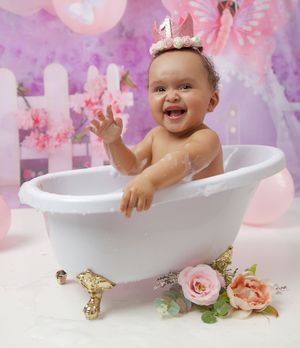 Baby sitting in a white clawfoot bathtub with pink roses and purple background for first birthday photoshoot.