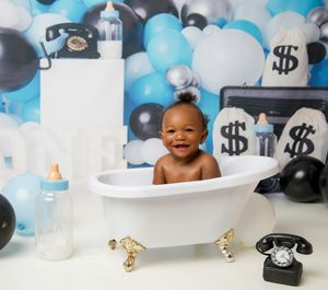 Baby in vintage clawfoot bathtub with blue and white balloon decor and retro phones in money-themed photo shoot setup.