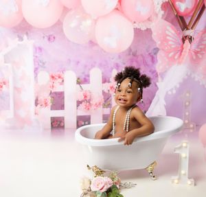 Pink balloon and butterfly themed bathroom photoshoot setup with clawfoot tub, flowers and white picket fence backdrop.