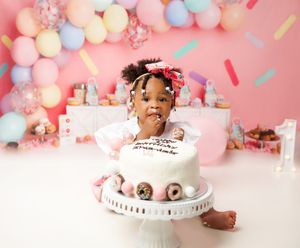 A festive first birthday cake smash setup with pastel balloons against a pink backdrop and white cake on pedestal.