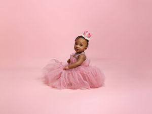 A small wearer in a fluffy pink tulle dress and crown sits gracefully against a pink backdrop during a studio photoshoot.
