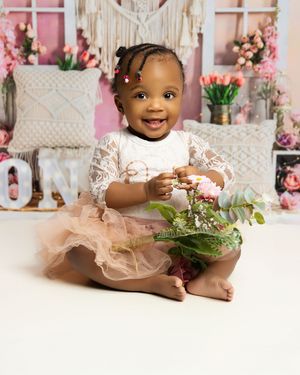Sweet one-year-old in lace dress and tulle skirt sits on floor with floral decorations and pink backdrop for birthday photos.