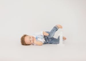 A toddler wearing jeans and a white shirt lies playfully on a white background.