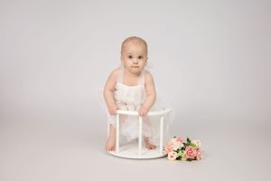 Small child in white outfit sits on round white stool next to pink floral bouquet on light gray background.