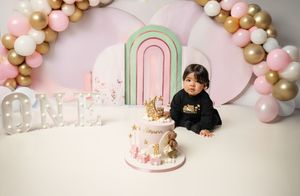 A pink and gold balloon arch decorates a first birthday party setup with a white ombre cake and pastel rainbow doorway backdrop.