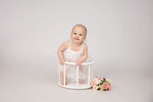 Baby in white outfit sits on small round white stool next to floral bouquet on light backdrop.