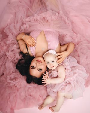 A mother and baby wearing matching pink tulle dresses lie together on a pink fluffy background.