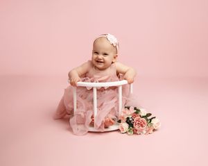 Baby in pink dress sits in white wooden stool prop with pink floral arrangement on pink backdrop.