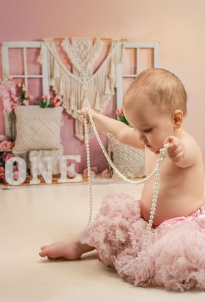 Baby wearing pink tutu and playing with pearl necklace in front of ONE letters and macrame wall decor backdrop.