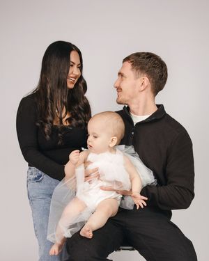 A couple wearing dark tops shares a loving moment with their baby in white against a light gray studio background.