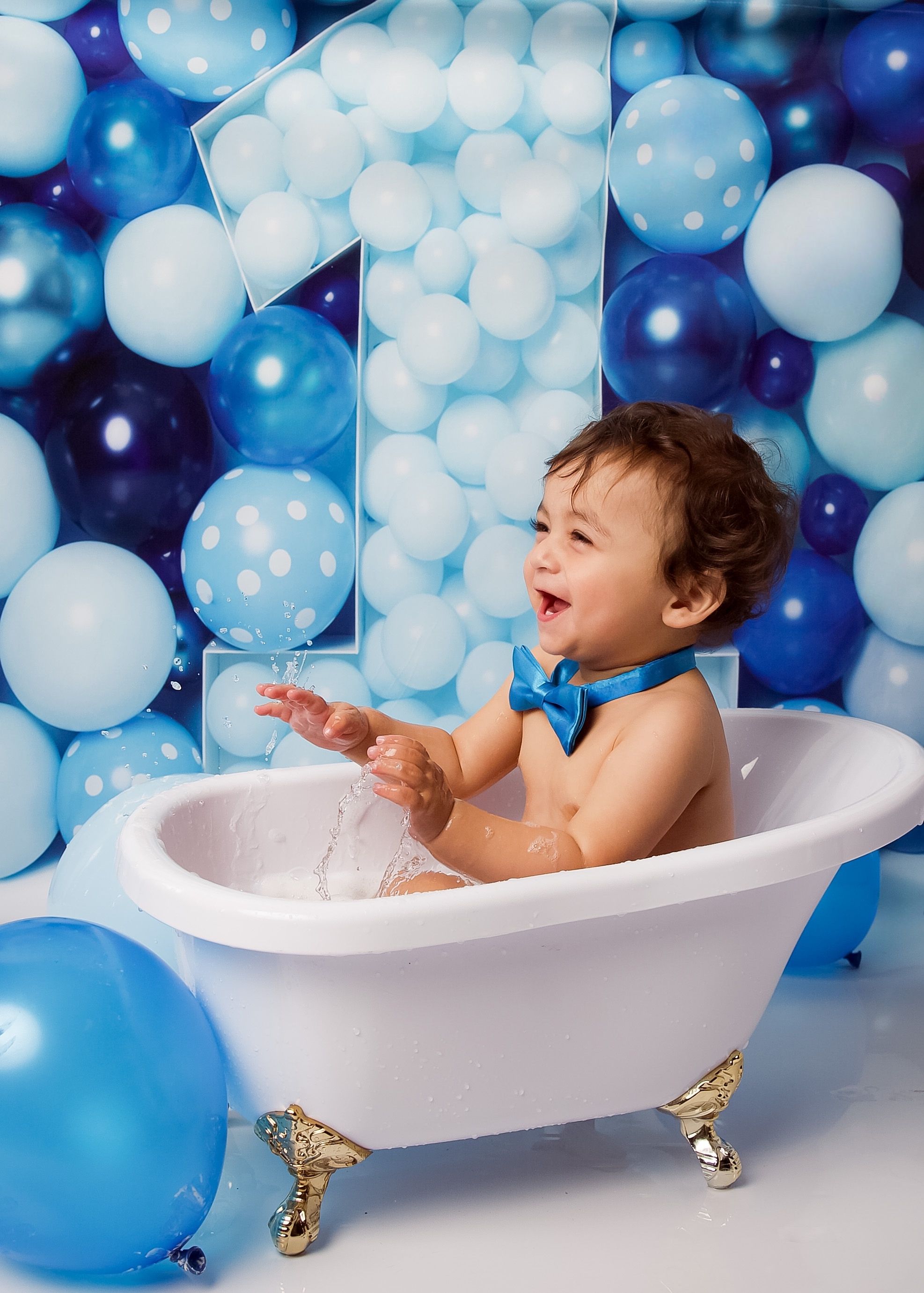 Joyful child plays in white clawfoot bathtub surrounded by blue and white balloons for first birthday photoshoot.