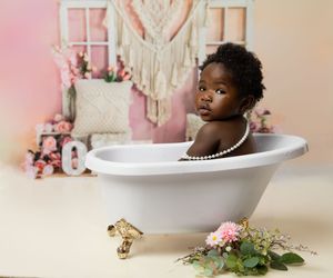 A young child sits in a vintage white clawfoot bathtub styled with pearls, macrame and pink flowers in a dreamy setting.