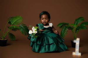 First birthday photoshoot featuring an emerald green satin dress with white flowers and tropical plants against brown backdrop.