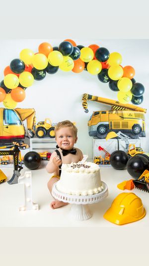 Construction themed birthday celebration with yellow and black balloon arch, toy trucks, and white cake on pedestal stand.