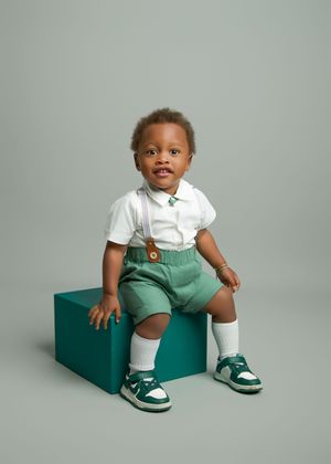 Young child in a classic preppy outfit with white shirt, green shorts and matching green sneakers sits on a teal cube pedestal.