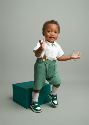 Small child wearing a white shirt and green shorts poses expressively while standing next to a green cube in retro-style shoes.