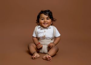 Baby wearing a white romper sits on brown backdrop during professional studio photoshoot.