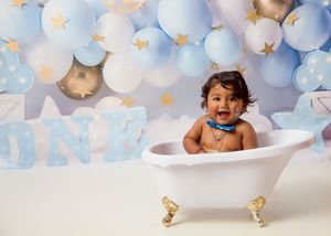 Baby sitting in white clawfoot bathtub with blue and white balloon decorations and star accents in background.