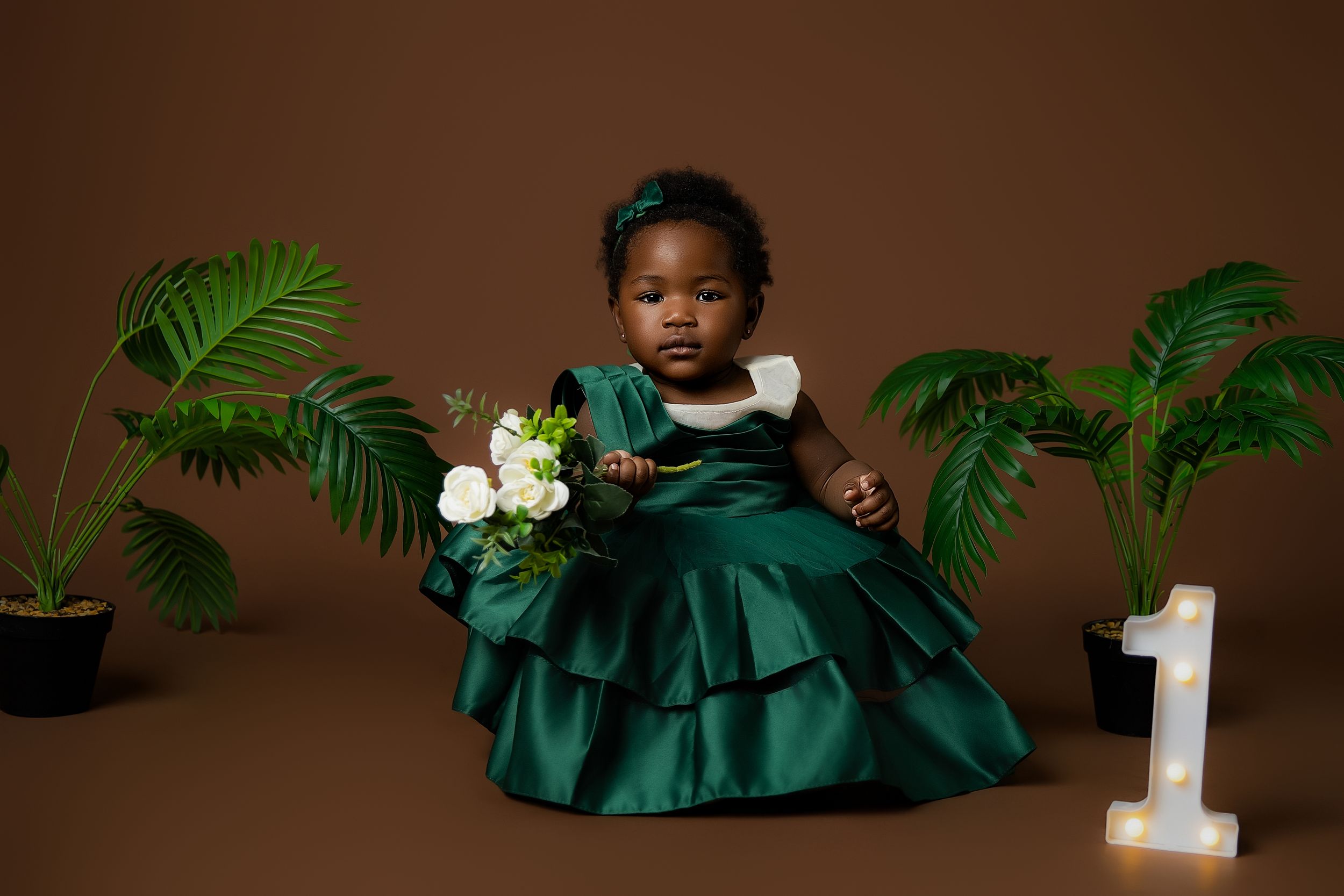 First birthday portrait featuring an elegant emerald green dress with white flowers and tropical plants against a brown backdrop.