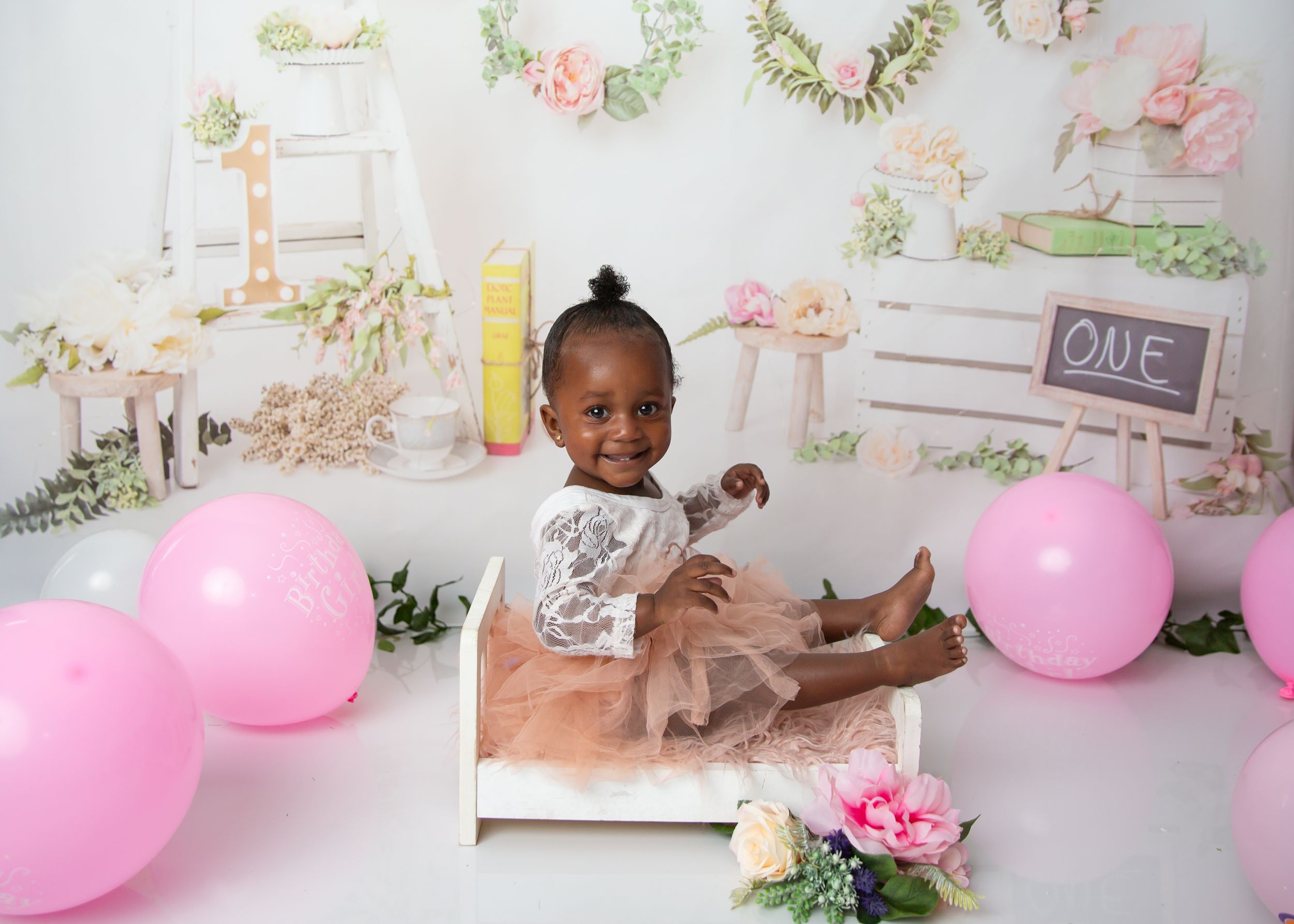 A one-year-old child posed in a pink and floral photo set for a first birthday cake smash session.